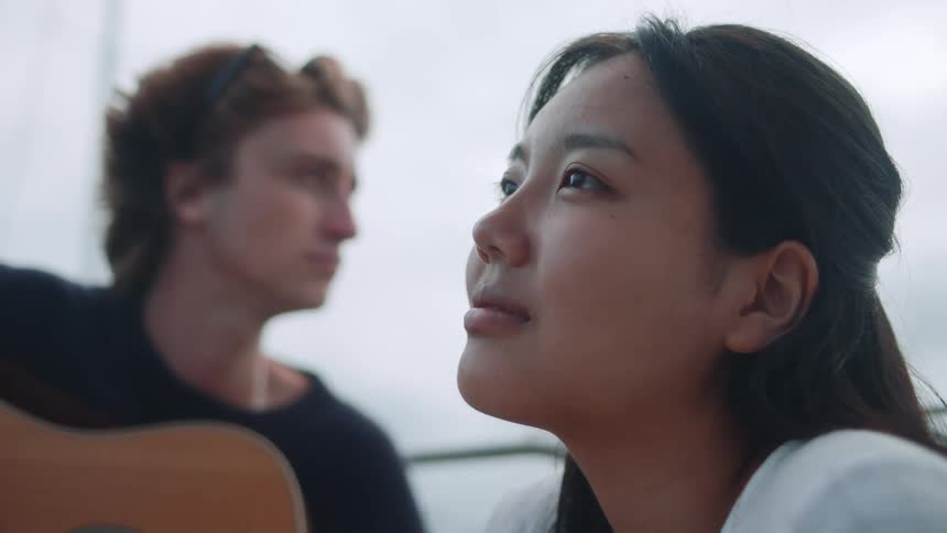 Woman smiles slightly as she and man with guitar sit on boat