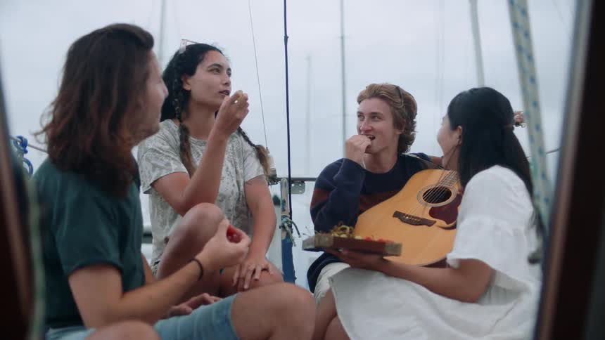 Group of friends relaxing on a sailboat eat fruit while talking at daytime