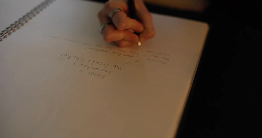 Woman with t shirt and black pants sits on couch and writes down in notebook in the living room