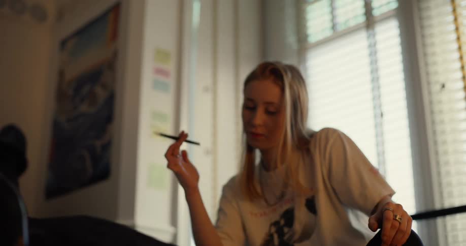Blonde woman with necklace and white t shirt thinks about an idea and writes down in notebook in living room in front of windows with blinds