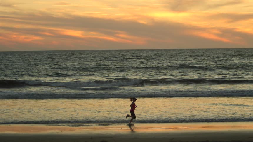 A woman is running along the beach through water at sunrise while waves roll in