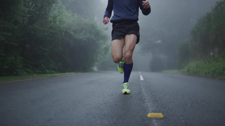 Man with blue shirt black shorts white and blue socks and sneakers jogs along the road through green forest in fog