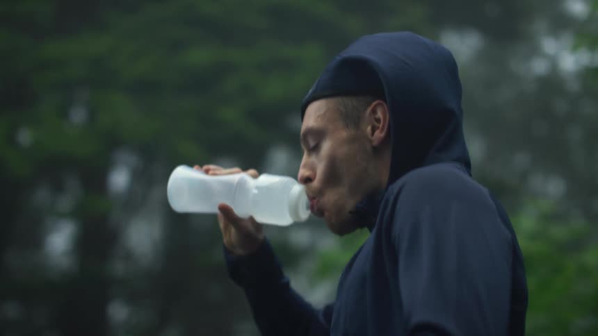 A man wearing a dark hoodie is jogging on a road near trees as he drinks from a water bottle