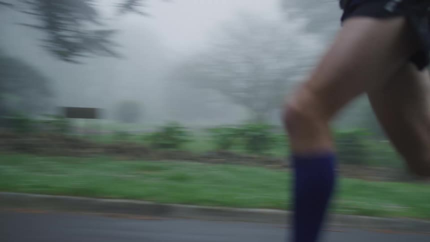 A jogger wearing shorts and a dark hoodie is running on a foggy rural road surrounded by trees