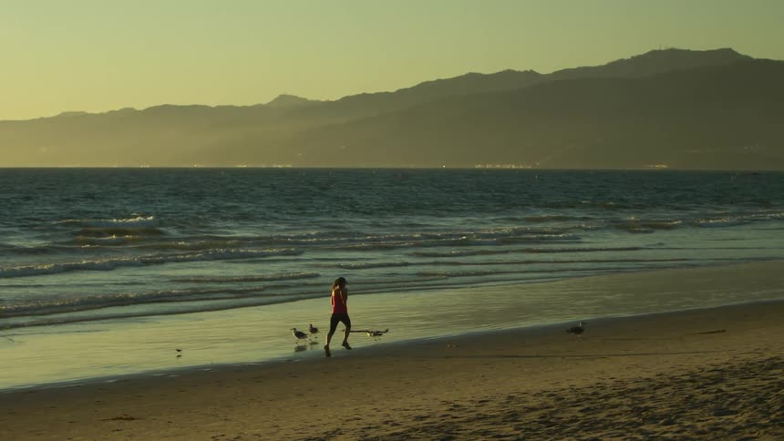 Woman with red sports shirt and shorts runs along beach near the ocean at sunrise