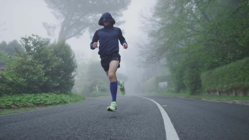 A jogger wearing shorts and a dark hoodie is running on a foggy road