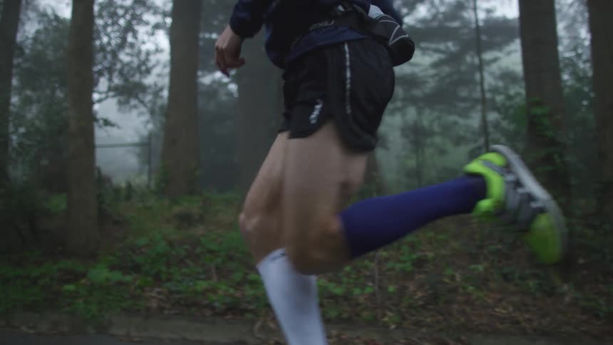 A jogger wearing shorts and a dark hoodie is running by trees on a foggy road