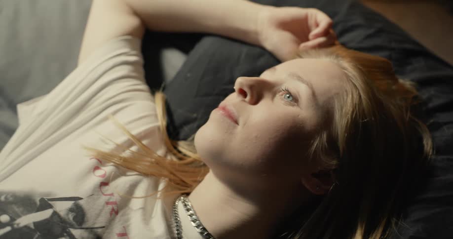 Blonde woman with necklace and white shirt lies on bed thinking and smiling