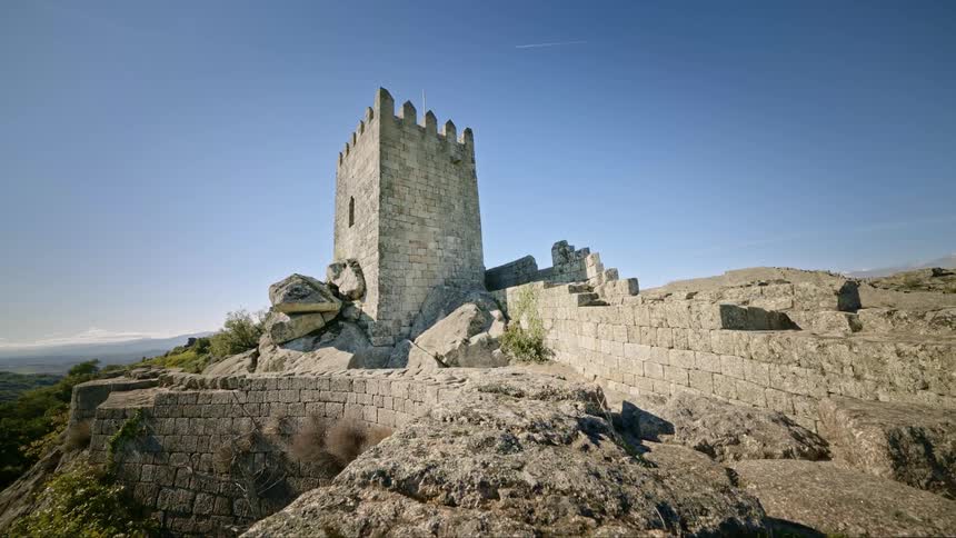 An old castle with a surrounding wall sits on top of a mountain under a clear sky