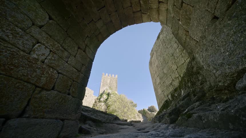 Looking out from the ruins of an old building and a wall a tower stands tall in a clear blue sky
