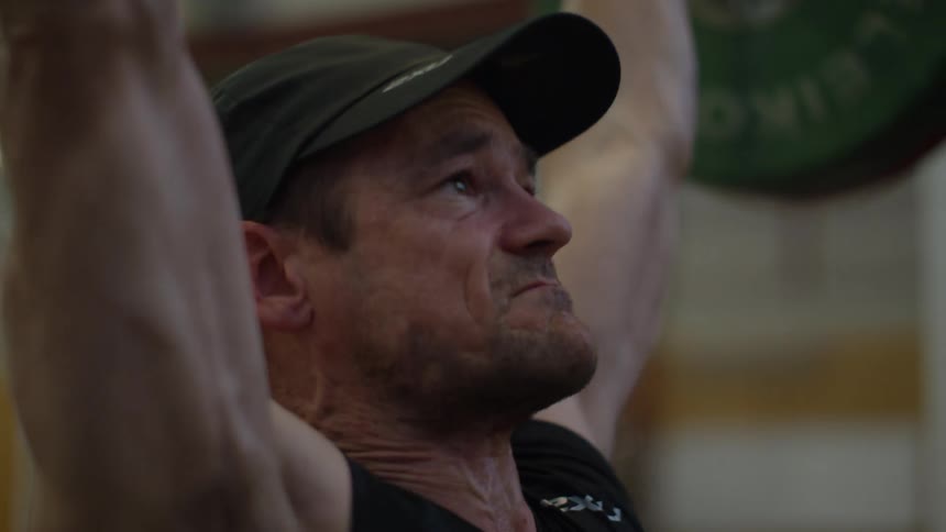 Man with cap and black shirt lifts up heavy barbell in the gym