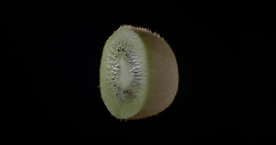 Vibrant green slice of fresh organic kiwi fruit is turning in the air against a black backdrop