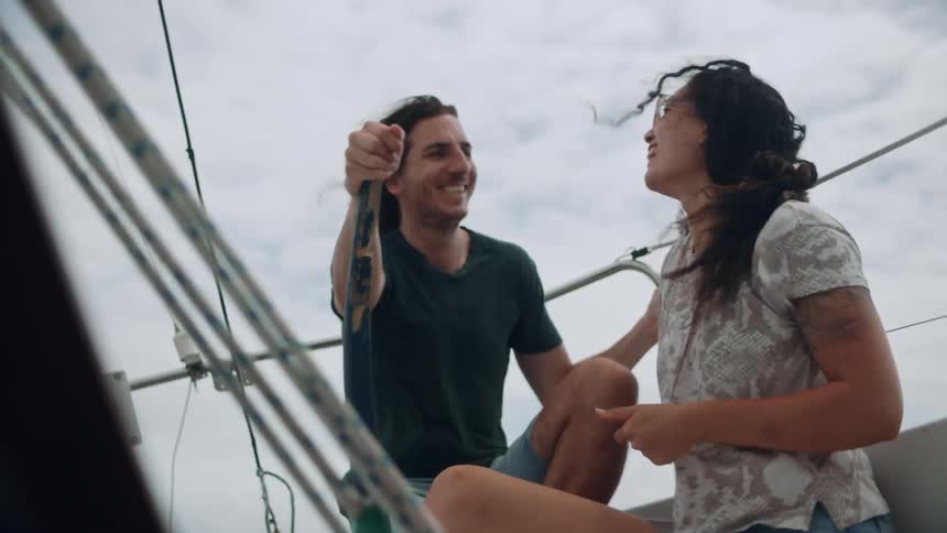 Man and woman in sailboat smile as boat bobs under overcast sky on windy day