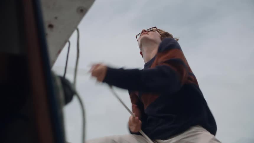 Young long haired man wearing sunglasses energetically pulls ropes on a sailboat