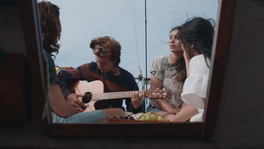 Friends sit on sailboat with fruit and listen to guitarist play