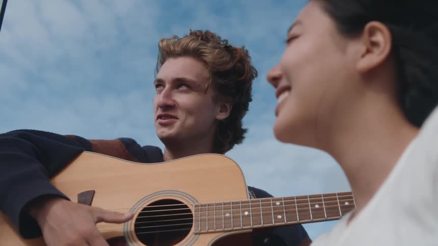 Man with black jacket plays guitar next to woman wearing white shirt while traveling on boat