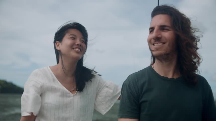 Man and woman smile while standing on sailboat passing rock outcropping