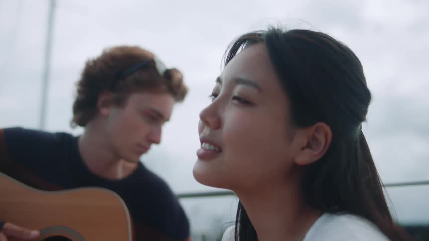 Girl with straight hair and white shirt looks up and smiles slightly while man playing guitar on boat on a cloudy day