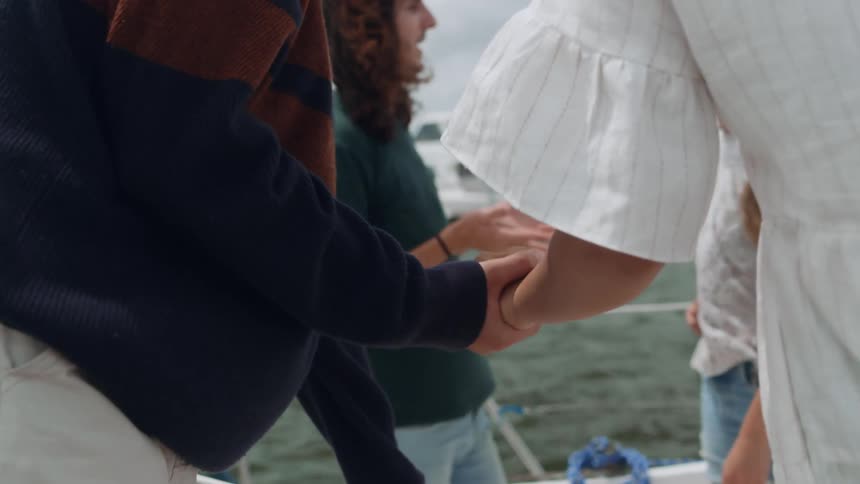 Male and female friends holding hands climb over the rail and join two other people chatting on a yacht in a bay on a sunny day
