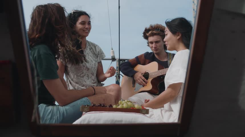 Friends sit on sailboat near fruit tray as one friend plays guitar
