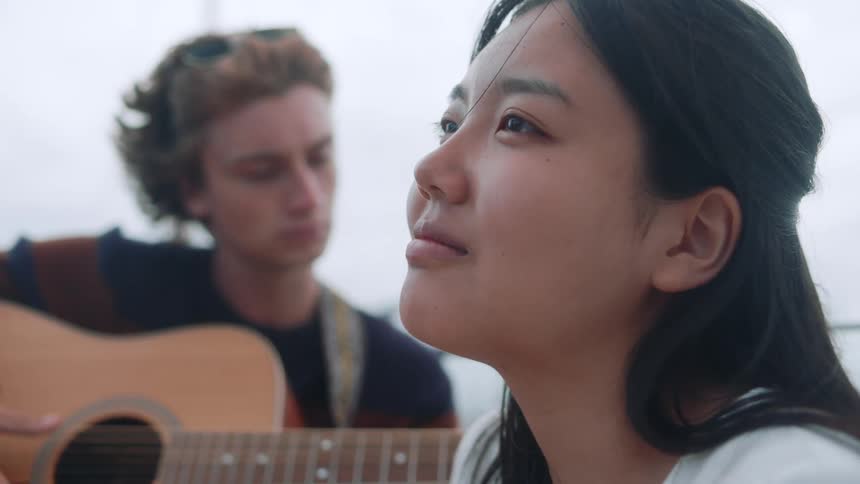 A man is playing a guitar while a woman is beside him and looking towards one direction with a happy expression on her face