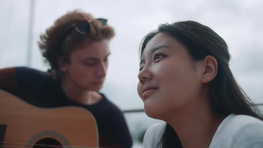 A guy looks down at the guitar he is holding than looks affectionately towards the girl sitting in front of him who is looking the other way dreamily while the boat they are on rocks gently