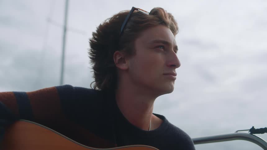Young man holding an acoustic guitar looks at the horizon while sitting on a boat