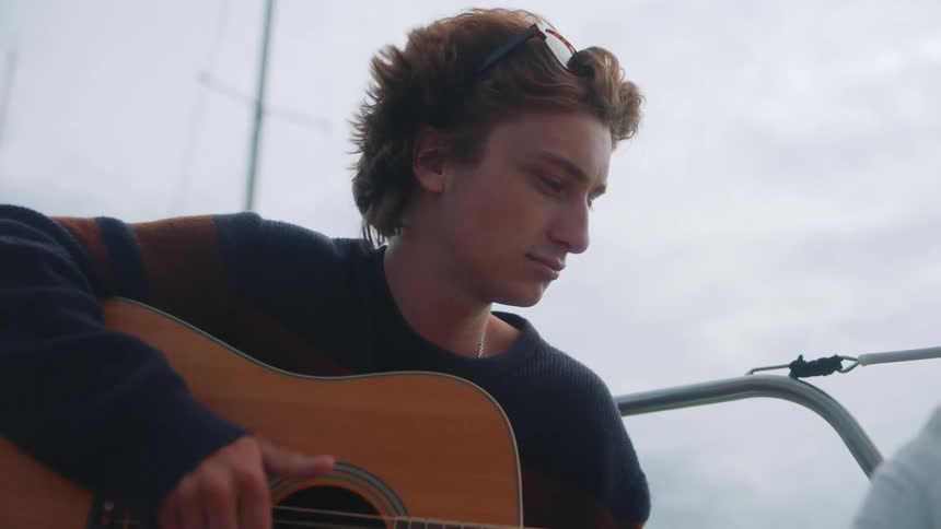 Man with sunglasses and blue sweater plays guitar on boat on a cloudy day