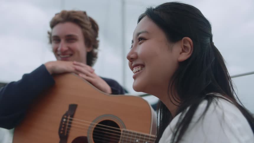 A teenage girl is smiling and laughing as a boy sits near her with an acoustic guitar in his lap