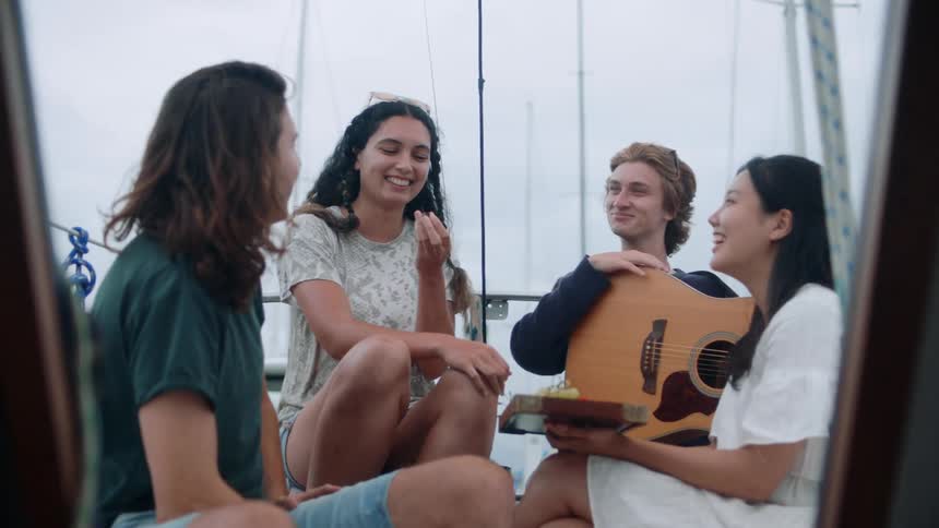 A group of friends two boys and two girls are sitting on a slightly rocking boat and talking and laughing while one of the males is holding a guitar and one of the females is holding a tray of the fruit the other female is eating from