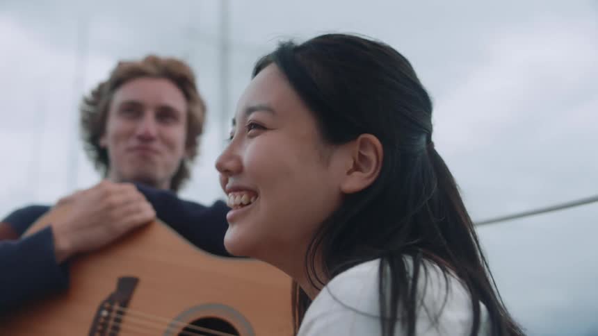 Man holding guitar and woman with straight hair talks to each other and laugh on the boat on a cloudy day