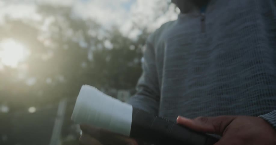 A black adult is wrapping a tennis racket with white tape while standing outdoors