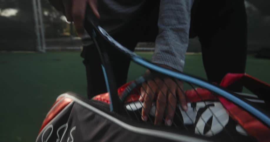 Man with gray shirt and black pants takes tennis racket out of red bag in tennis court