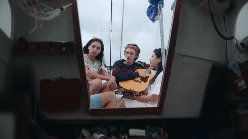 Four young people gathered on a sailboat eating talking playing guitar
