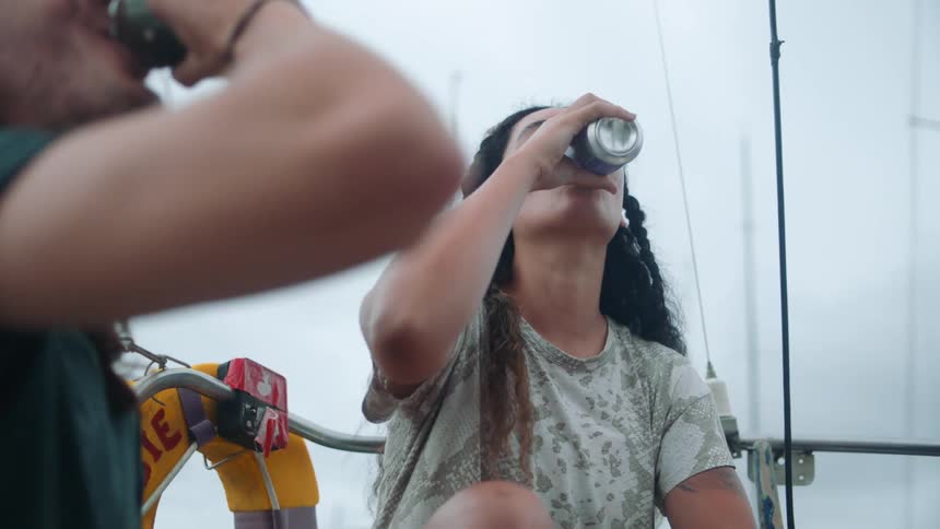 Two young happy friends toast with beer while sitting on a sailboat