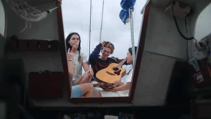 A group of friends are sitting on a boat talking and laughing while one of them is eating something and another is holding a guitar