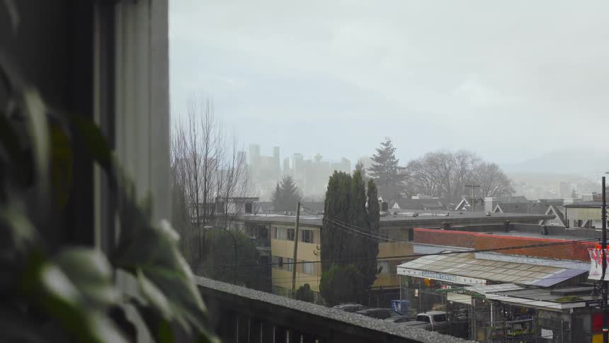 Hail is falling and landing on a balcony railing and the closely built apartment buildings below under a hazy white sky