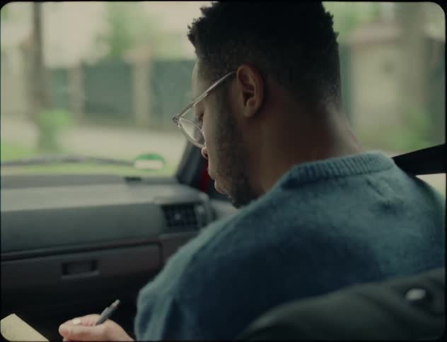 A black man is sitting in the passenger seat of a moving car and making a list while the driver makes quick hand gestures