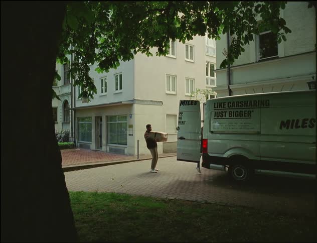 A man is standing behind a van parked near buildings as a woman jumps out with a box and hands it to him