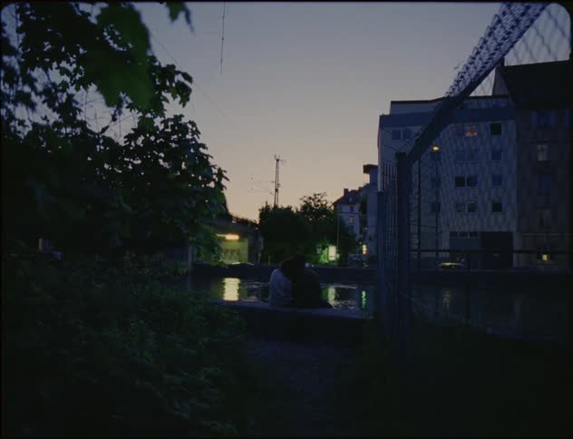 Couple cuddles while sitting on shore across river from apartment building at night