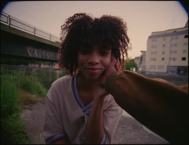 A woman is standing outside near a stream and a building with a bridge in the distance as a man reaches out and puts his hand on her face