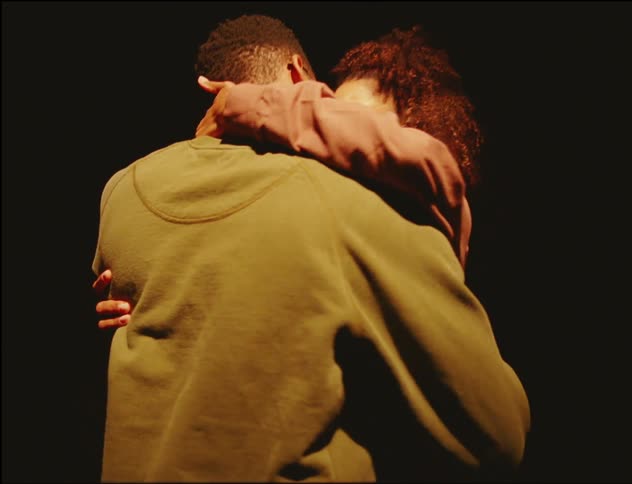 A man and a woman approach each other as everything else around them fades to black and they begin to embrace and kiss as they move closer than further away and a spotlight is on them