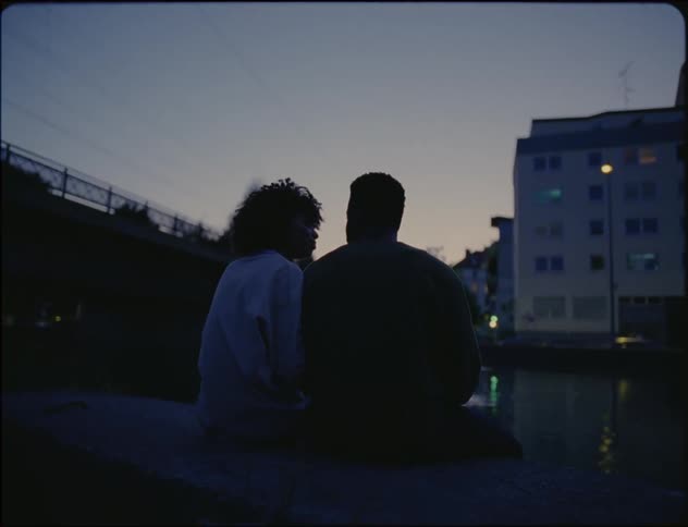 Couple sits on riverbank across from apartment building and kiss