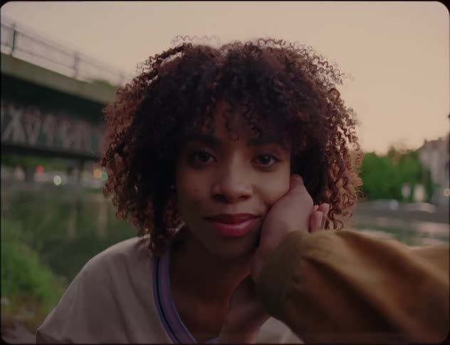 A woman is sitting outdoors looking forward fondly slightly smiling as a hand reaches out and touches her cheek gently as she puts her hand over it