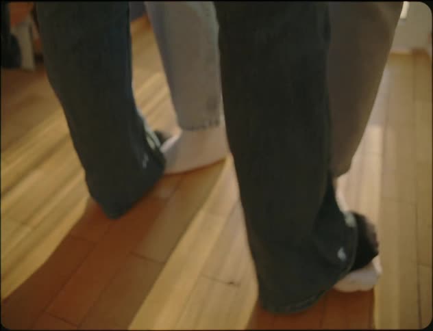 Two people are moving together in a dancing motion with one persons feet on top of the others in a room inside a home with wood floors