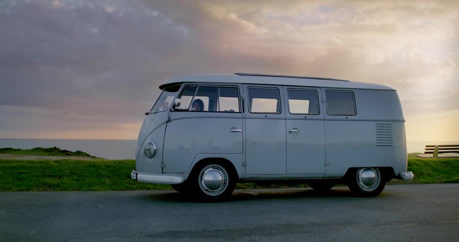 A woman is sitting in an old white van parked on a road overlooking the water