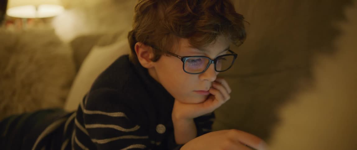 Young boy wearing glasses swipes on a tablet while laying on the couch in the living room