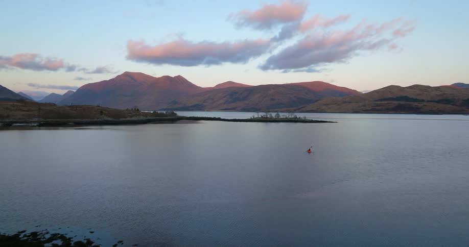 A small boat sails across the water as the person inside paddles toward land and there are mountains in the distance