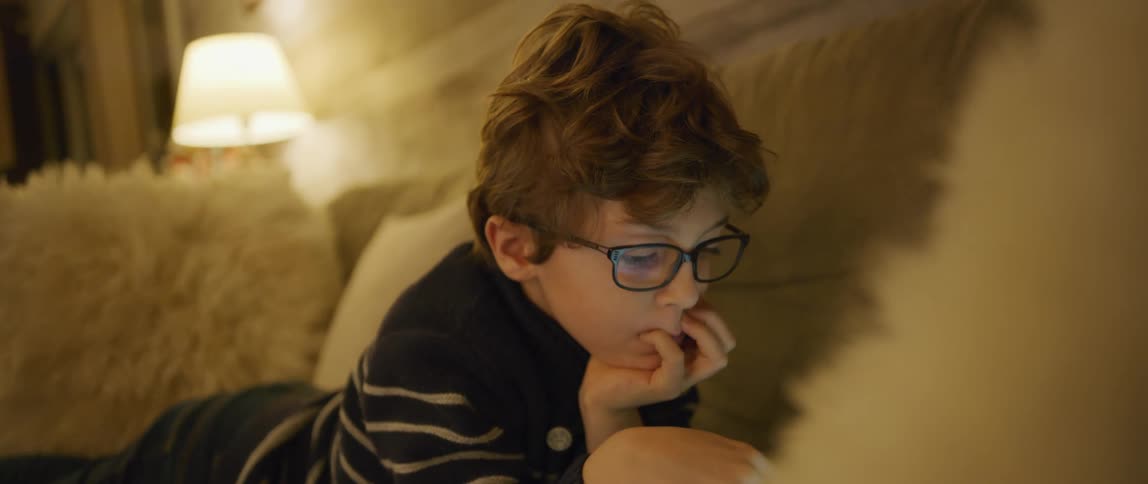 Boy wearing glasses plays with a tablet while laying on the living room floor