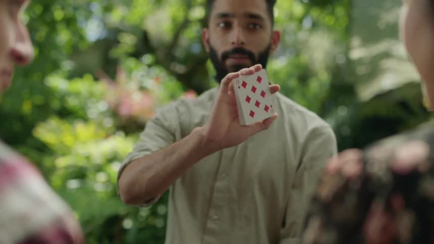 Young bearded magician entertains friends with card tricks in the garden